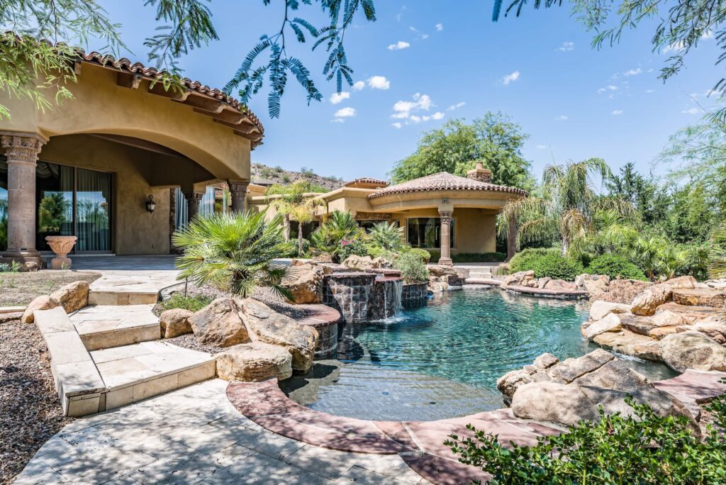 Luxury Arizona desert home with terracotta roof, large swimming pool with rock waterfall features, and lush landscaping.