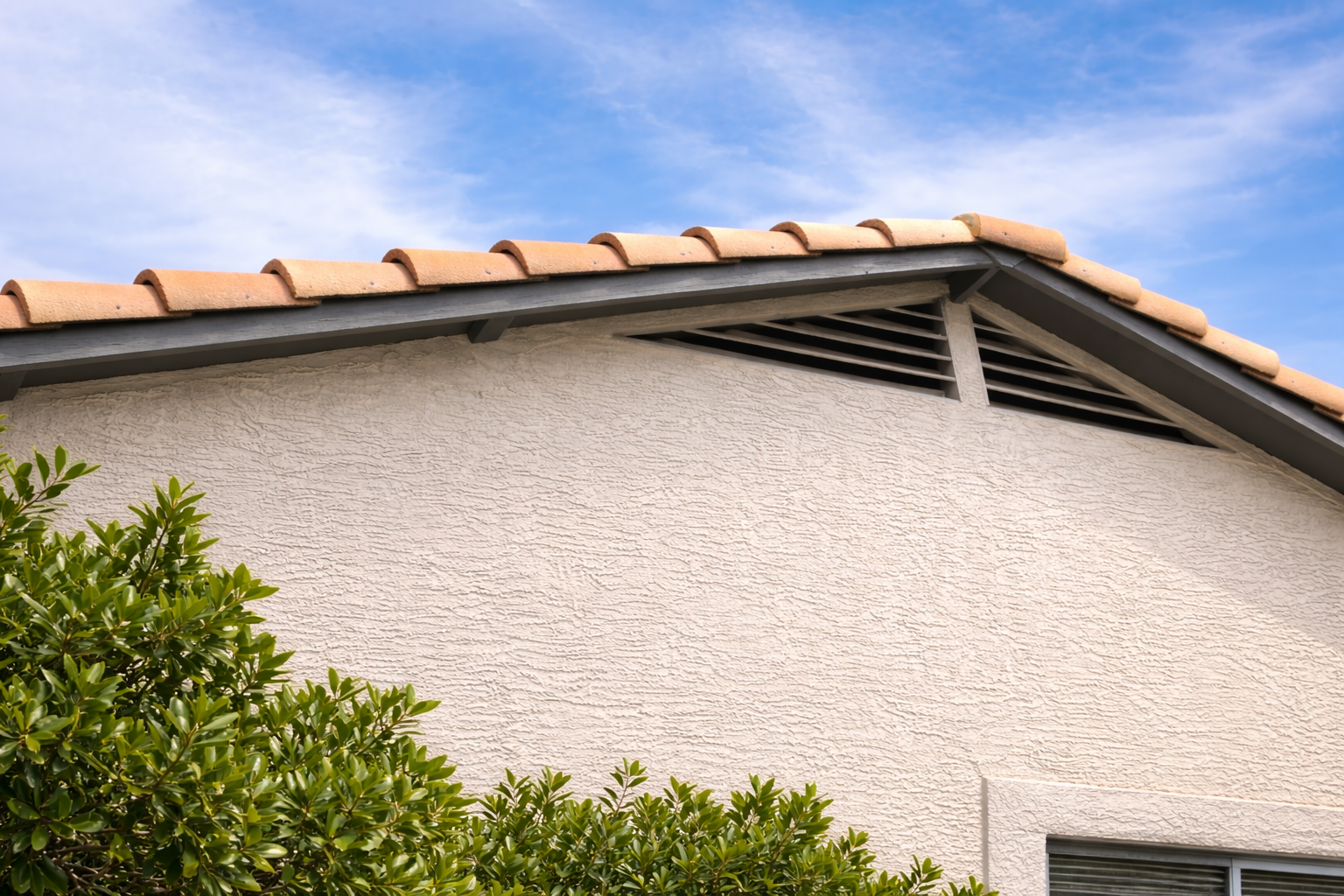 Terra cotta tile roof with a stucco wall and gable vent on a house in Glendale, Arizona, with green bushes.